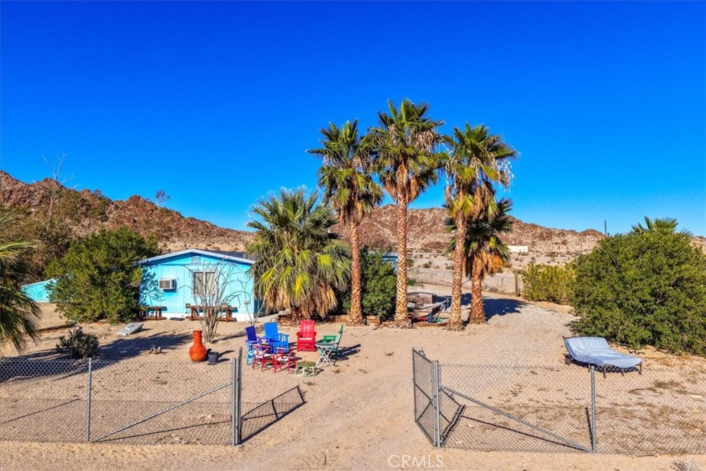 Walk beneath the palms and feel the world quiet. Step through the gate of Blue Desert Oasis and you cross a threshold—leaving behind the open Wonder Valley expanse for a space made lush by care and vision. Mature palms sway above desert blooms, the air cool beneath their shade. The home sits on five fully fenced acres, its own private well sustaining this tranquil retreat near the base of the Valley Mountains. The shaded front porch sets the pace—slow, grounded, welcoming. Inside, the main living area opens wide with tile floors, generous windows, and clean desert light. The kitchen carries a handcrafted warmth with Talavera tile counters and rich wooden cabinetry, partially open to both the dining and living rooms. The bathroom features a walk-in shower and pedestal sink, and the expansive bedroom—large enough to feel almost like a studio of its own—offers outdoor access and ample closet space. Beyond the walls, the property continues to unfold: a detached two-car garage with laundry, two additional sheds (one powered), and carefully tended landscaping that softens the desert’s edges. From the yard, the view sweeps from the nearby Valley Mountains to the distant Pinto range of Joshua Tree National Park. Currently a beloved short-term rental, Blue Desert Oasis carries a history of arrival—each guest crossing that same quiet threshold into calm. Whether you keep it as a retreat for others or claim it as your own, this place waits patiently, ready to welcome whoever steps through the door next.