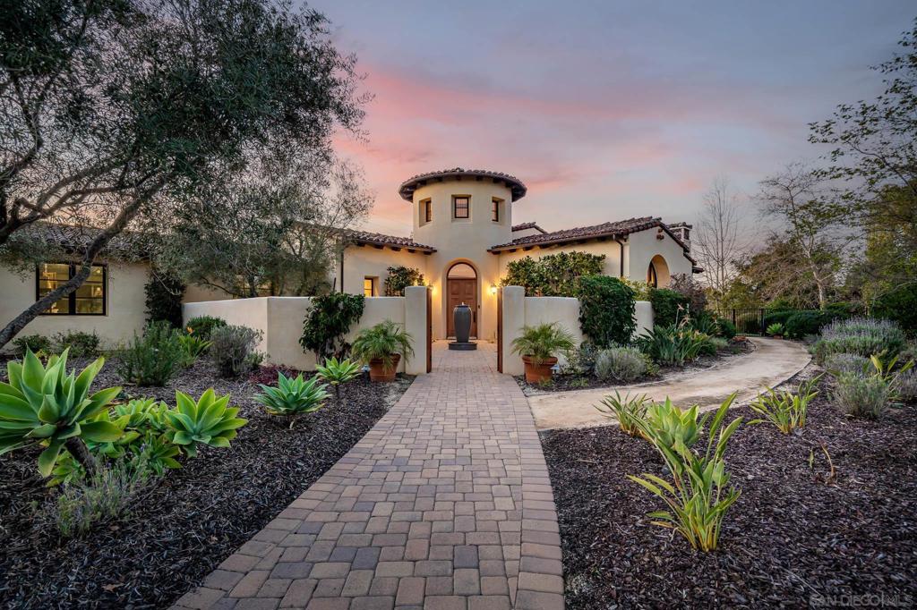 Welcome to 7552 Montien Road, a refined single-story estate built in 2014 by Mulvey Custom Builders and located in the prestigious guard-gated community of Santaluz. Designed to blur the line between indoors and out, this residence invites both effortless entertaining and quiet retreat. Through a serene entry courtyard, you’ll find a flowing open plan with soaring wood-beamed ceilings, travertine floors, and curated designer finishes. The chef’s kitchen combines beauty and function with custom cabinetry, GE Monogram appliances, dual ovens and dishwashers, a 72” built-in refrigerator, 6-burner cooktop, and pot filler—all oriented toward sweeping views of the pool and mountains. The primary suite is a true haven, featuring a spa-like bath with steam shower, soaking tub, fireplace, and French doors that open to a private patio. Each secondary bedroom is generously scaled with walk-in closets and en-suite baths. A tiered theater with pocket doors provides a cinematic experience at home. Outdoors, a resort-style pool and spa, built-in bar with BBQ, and expansive patios offer endless enjoyment under San Diego skies atop a 1.74 acre lot. Set on one of Santaluz’s most sought-after parcels overlooking the 7th green of the Rees Jones-designed course, this residence also benefits from the community’s 3,800 acres of rolling hills, 33 miles of hiking and biking trails, and two clubhouses. Membership opportunities include golf, tennis, spa, fitness, dining, and social events, creating a lifestyle that is both active and secure.