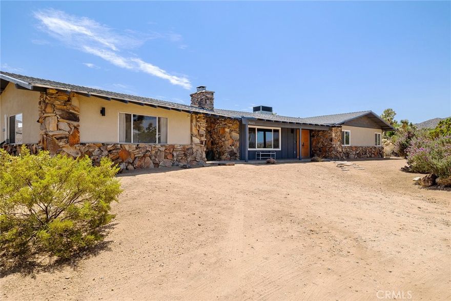 Classic Midcentury Charm, Floor plan and Architecture with sunken living room. Midcentury charm starts with curb appeal, this home is set back from the street, centered on a square 1.4 acre lot. A long-looped driveway through lush hi-desert plants and bushes including large Joshua Trees and a large rear facing garage, a classic feature of Yucca Valley homes because of their large lots and relaxing lifestyle, most commonly built by early developer Fred A. Storey.  The face of the home features rock masonry, exposed beams and eaves, a welcoming front patio and breezeway. The entry features double doors, paneled and carved. The entry looks on to a sunken living room, a giant window with beamed ceilings and a grand rock fireplace. The home has polished concrete floors throughout. To the right a den/office/bedroom with an ornate door. Forward through a pocket door is an open concept kitchen, dining room and laundry room with doors to the yard and breezeway. The bedroom wing hall has a large bank of built cabinets for extra storage. The guest bathroom is large and remodeled with terrazzo tile floors, floating vanity and a walk-in shower. The guest bedroom is large, with a large closet and windows. The primary bedroom is very large, with a walk-in closet, and remodeled full bath. The yard is big, the amenities spaced out with large Joshua Trees and pathways and wide views of Joshua Tree National Park Mountains. The large rear facing garage with its huge glass door and finished game room interior and mini split for heating and cool it’s the perfect place to enjoy the view and preside over a gathering enjoying the pool, spa, sauna and sundeck.