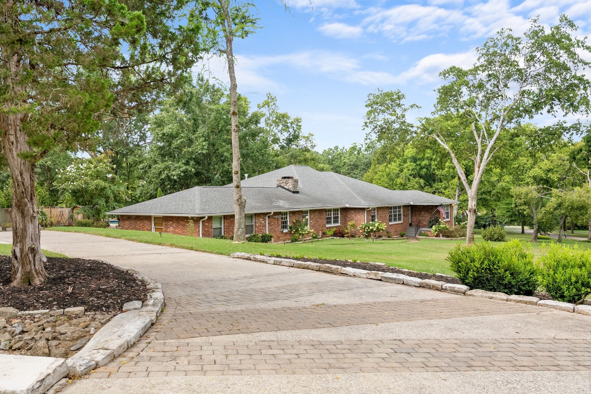 3-bed home for sale in Nashville, TN 37205, 615 W Meade Dr Long brick driveway with a paver surface leading to the front of a single-story brick house with a gray shingle roof and a stone chimney