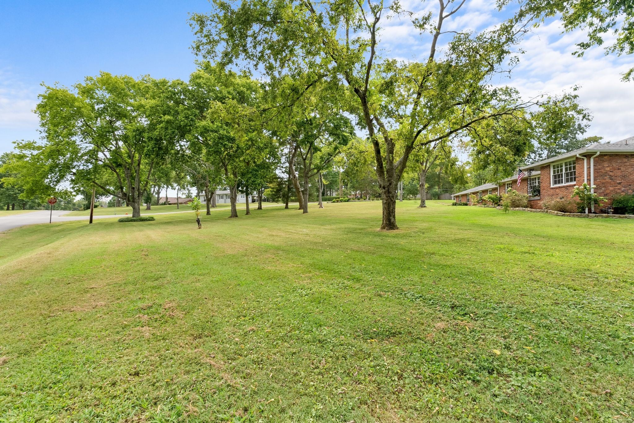 3-bed home for sale in Nashville, TN 37205, 615 W Meade Dr Open grassy yard lined with tall trees and a brick side wall of the house on the right with white-framed windows