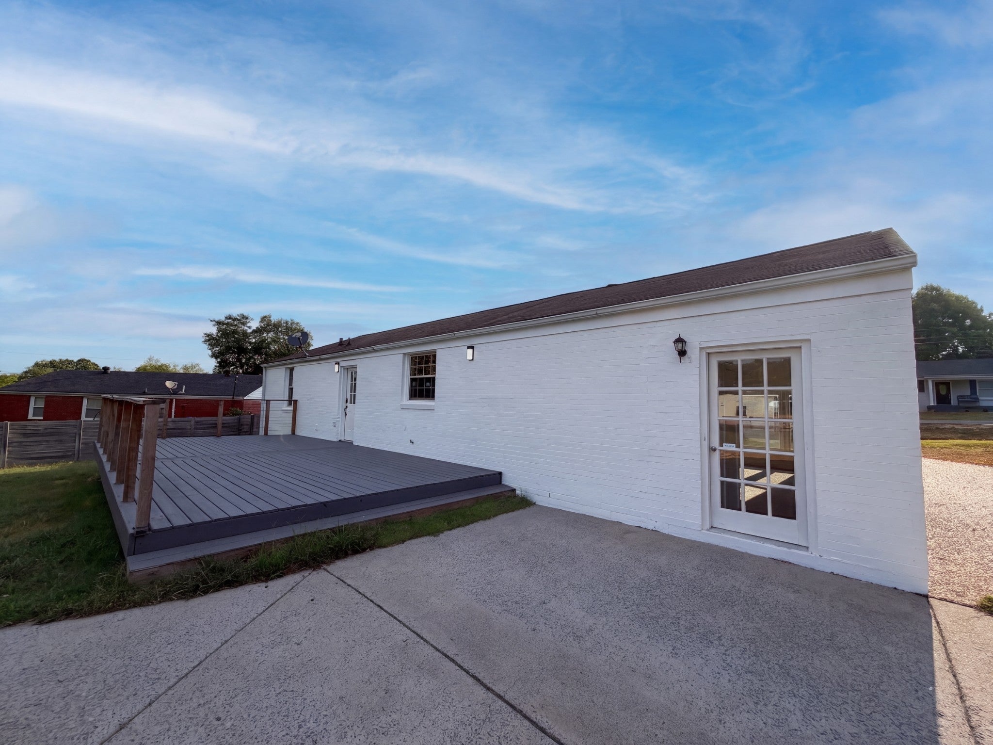Rear of the house with white brick walls, a gray wood deck on the left, a glass-paneled door and a concrete patio