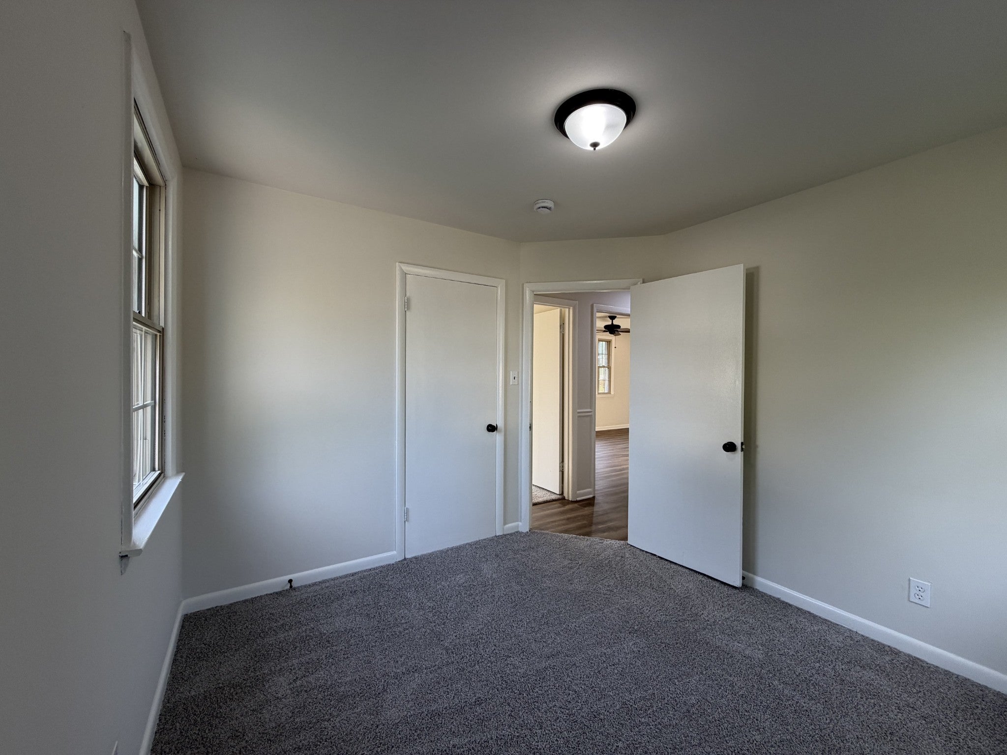 Bedroom with gray carpet, a ceiling light, a window and an open door leading to the hallway