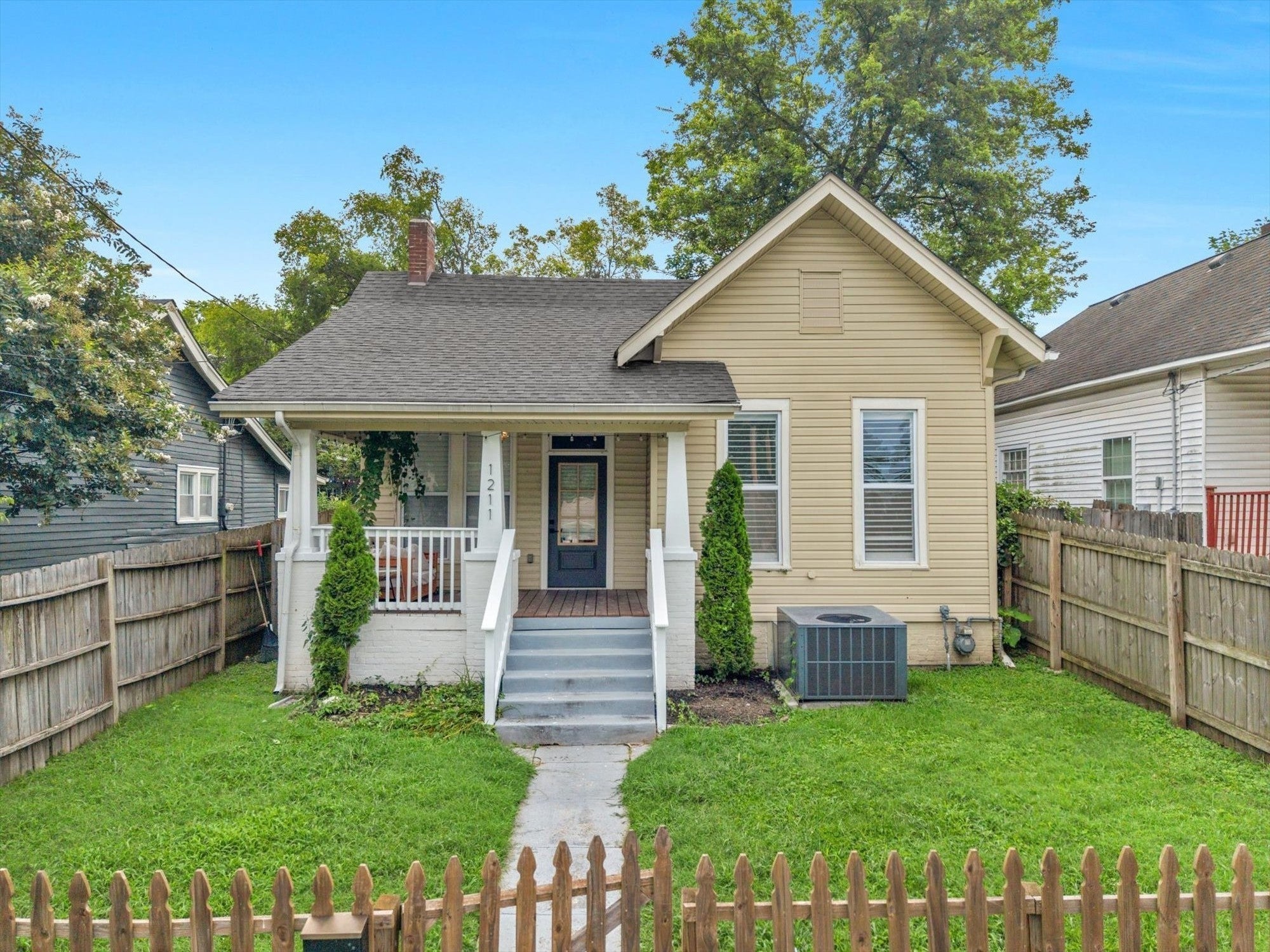 3-bed home for sale in Nashville, TN 37207, 1211 Stainback Ave Front exterior of a tan house with a porch, white railings, a side yard lawn and a wood privacy fence along the lot
