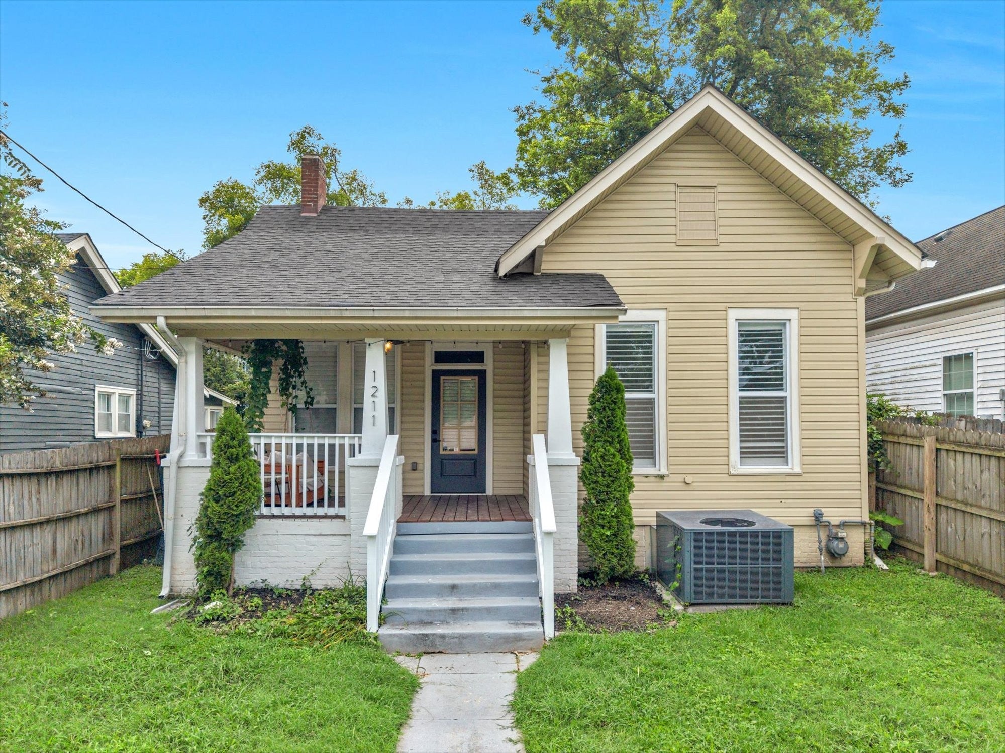 3-bed home for sale in Nashville, TN 37207, 1211 Stainback Ave Front view of a tan single-story house with a gabled roof, a covered porch, concrete steps and a side yard lawn
