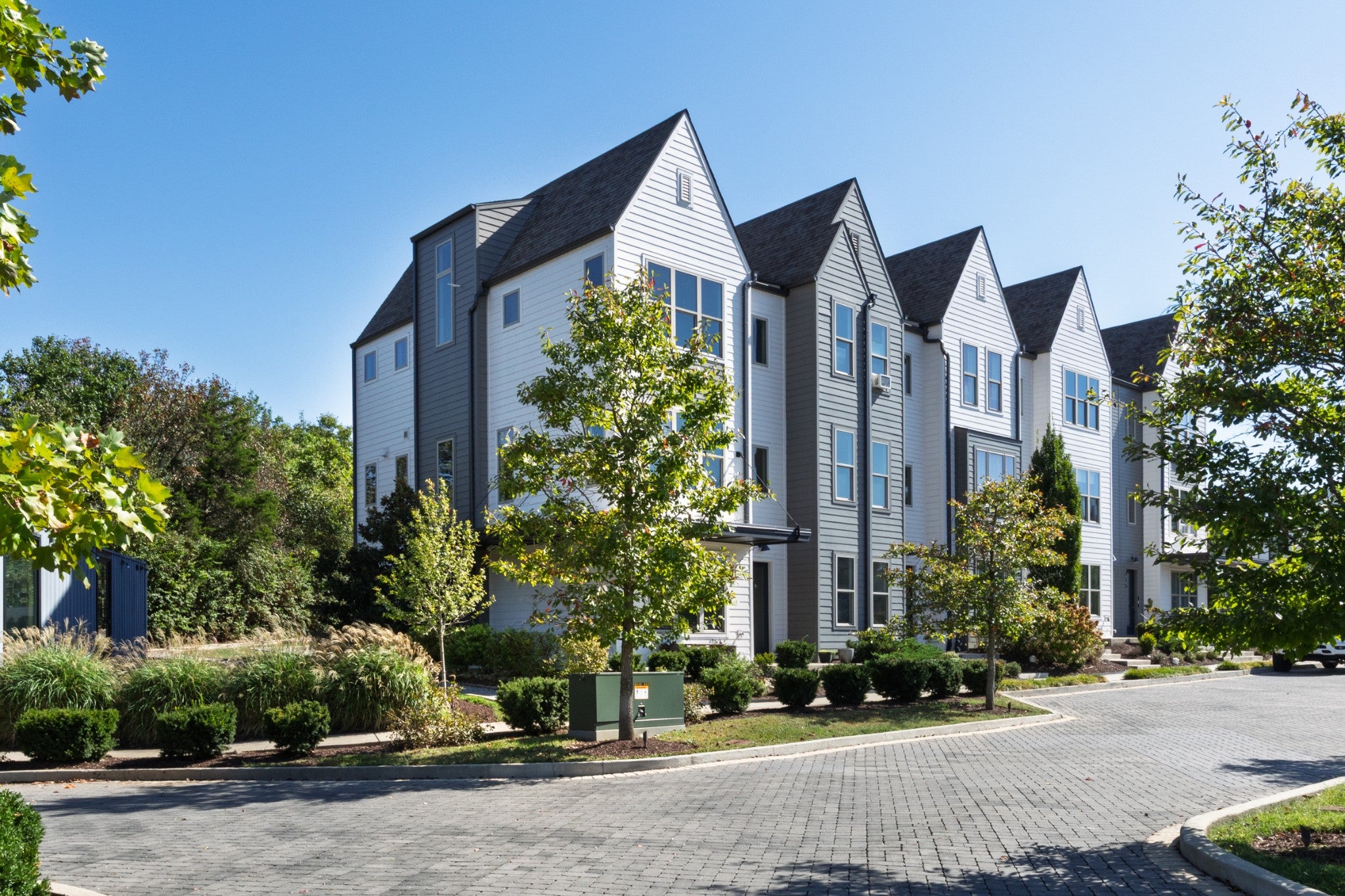 Row of attached townhomes with gabled roofs and gray and white siding, lined by a brick paver drive and low shrubs