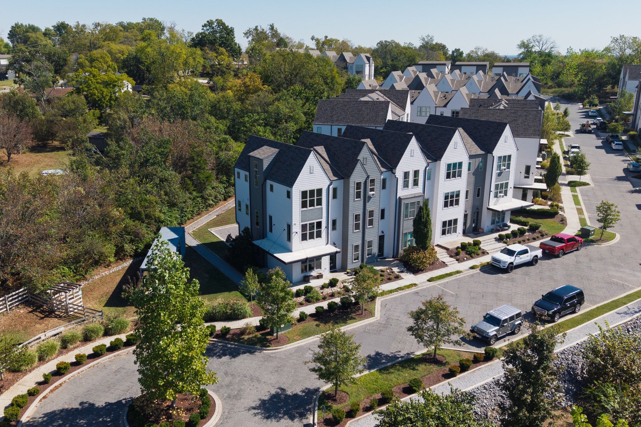 Aerial view of townhomes on a curved street with parked vehicles, trees and a wooded lot beside the row