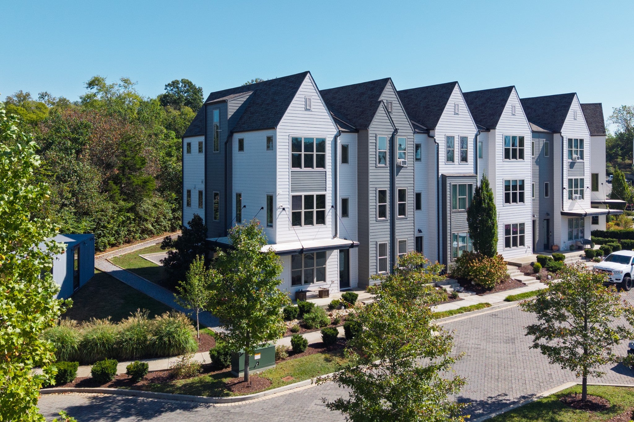 Aerial view of a row of three-story townhomes with dark shingle roofs, paver parking and a metal utility box in the front yard