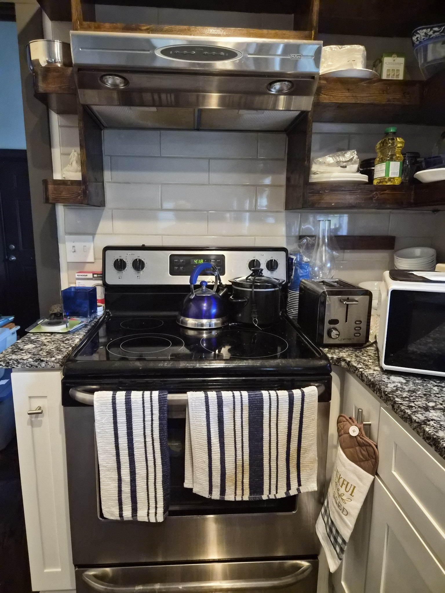 Kitchen range with a stainless steel hood, white subway tile backsplash and wood shelves above the cooktop