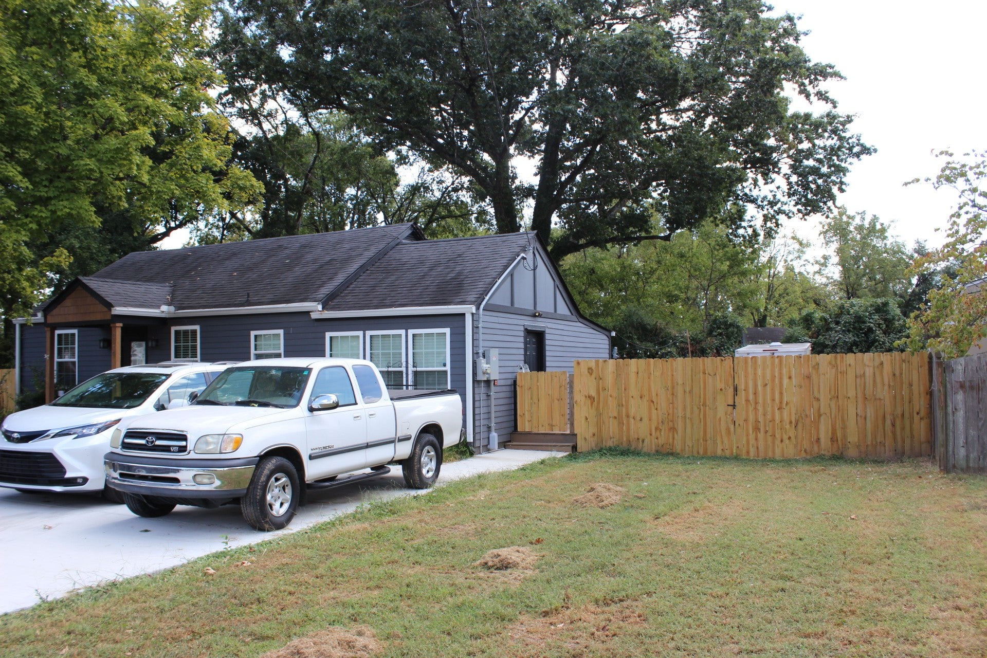 Side view of the house with gray siding, a white SUV and a white pickup in the concrete drive, and a tall wood privacy fence
