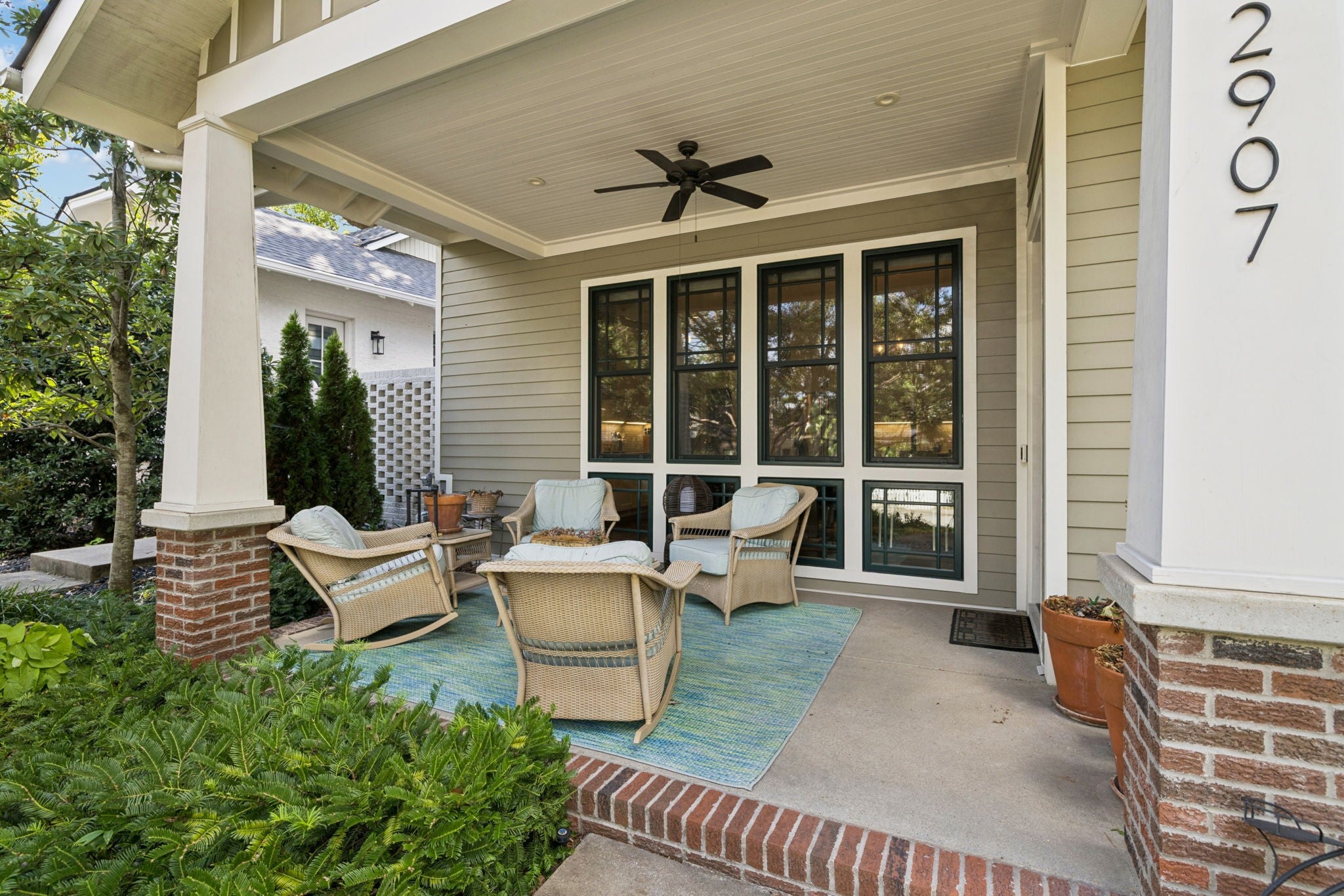 Covered front porch with wicker chairs on a blue rug, a ceiling fan and tall windows beside the entry