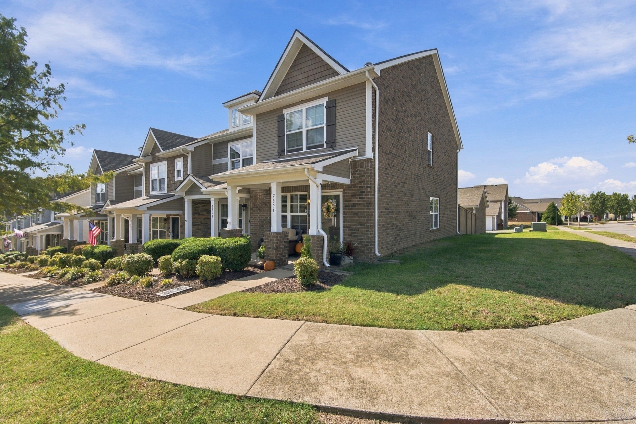 Side view of a two-story brick and tan siding townhome with a covered porch and a lawn along a sidewalk corner