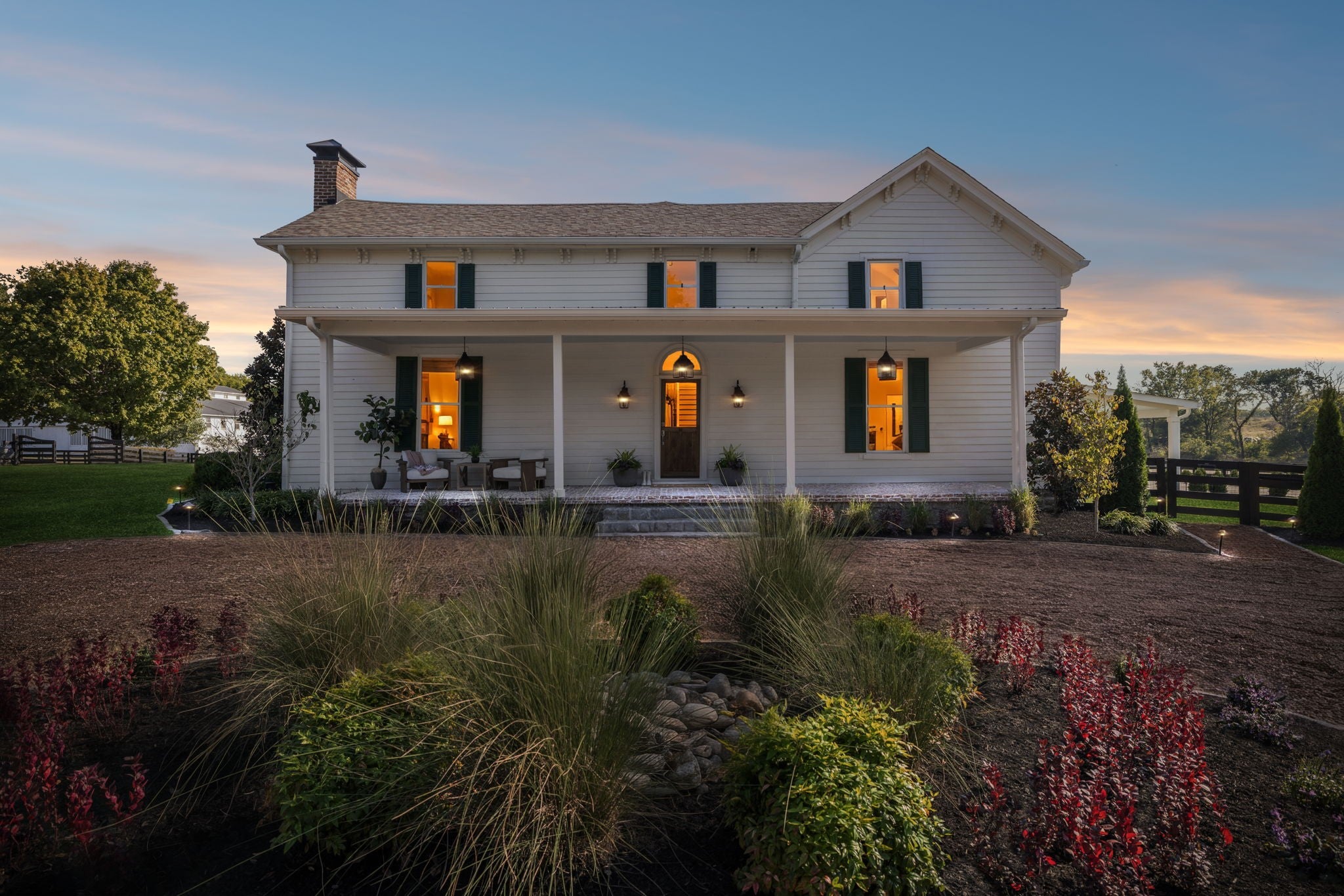 4-bed home for sale in College Grove, TN 37046, 6615 Eudailey Covington Rd Two-story white farmhouse with a front porch, green shutters and warm lights glowing at dusk, with plantings along the front walk