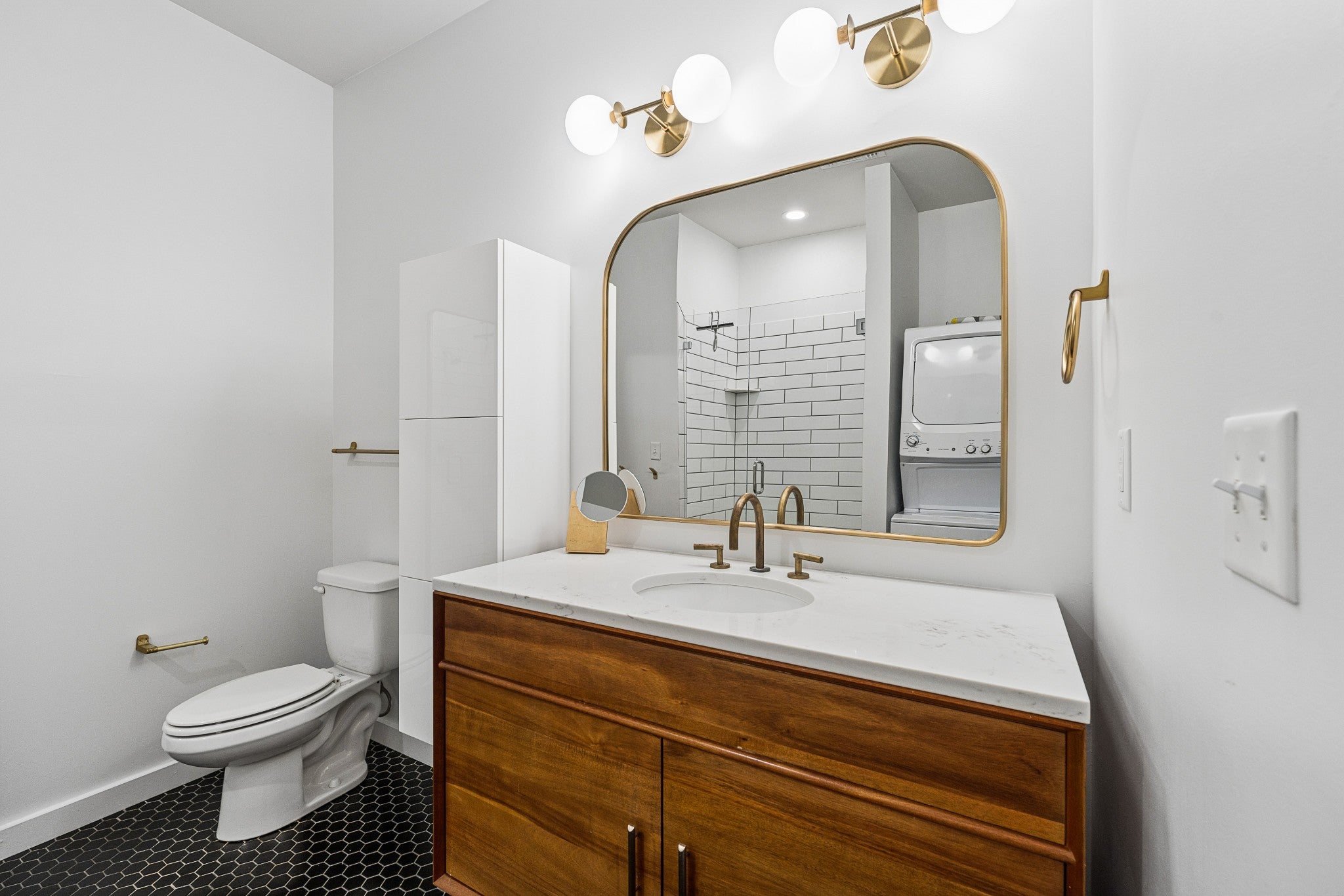 Bathroom with a wood vanity, white countertop, black hexagon floor tile and a tiled shower beside a stacked washer and dryer