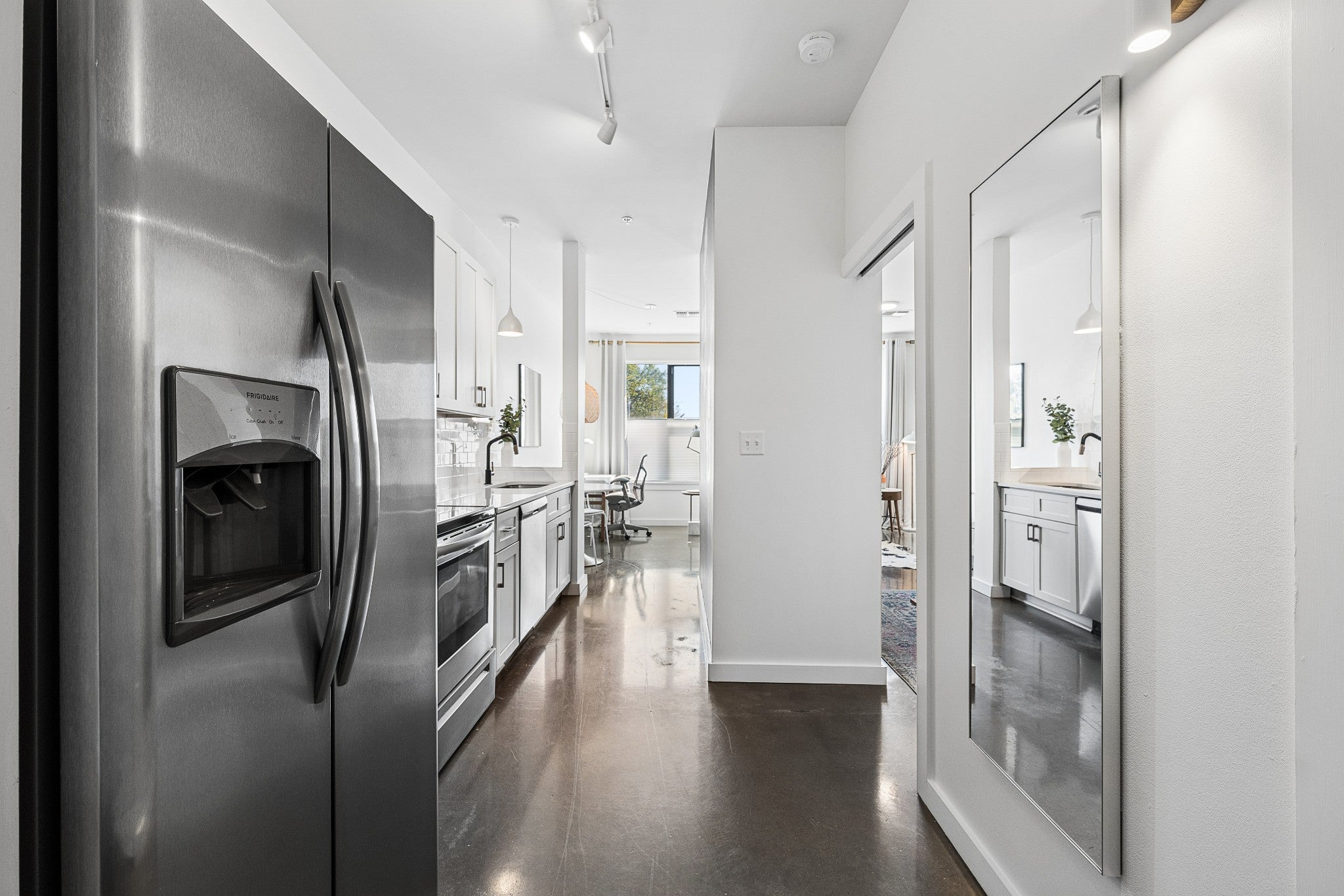 Hallway with stainless steel refrigerator, white cabinets, track lighting and a wall mirror leading toward the living area