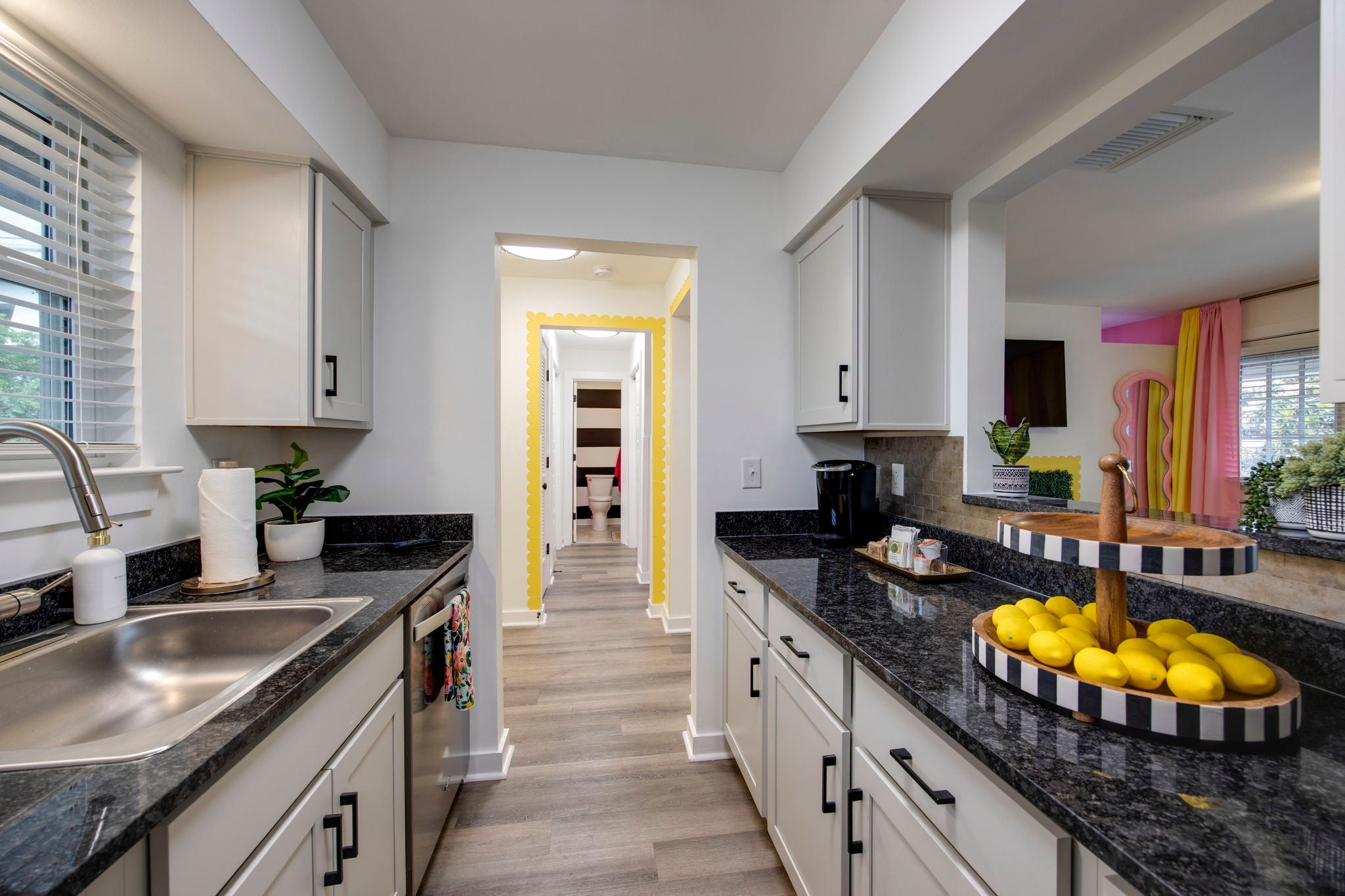 Galley kitchen with white cabinets, dark stone countertops, a stainless steel sink and a yellow-trimmed hallway ahead