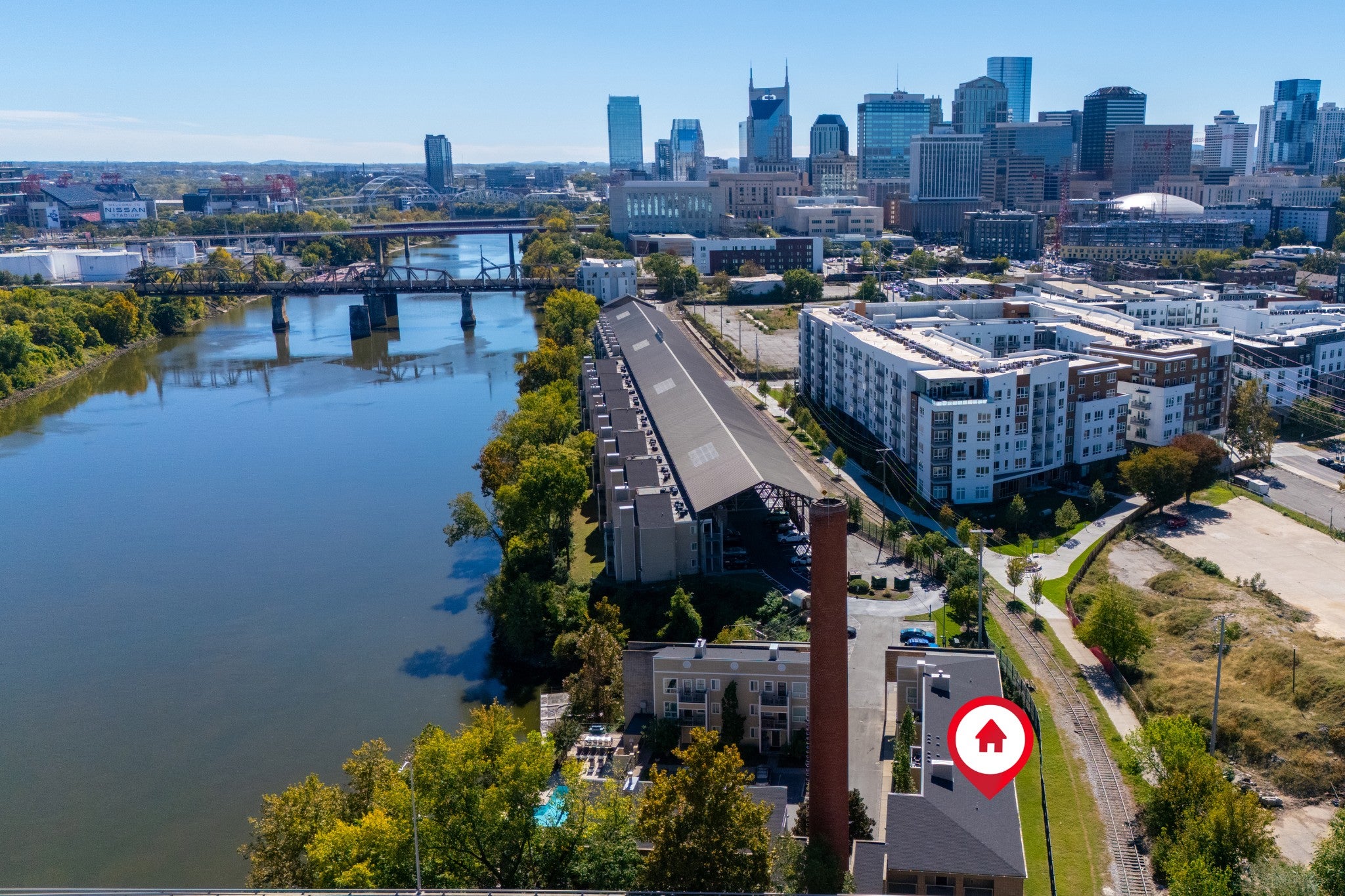 Aerial view of a riverside building with a brick chimney, a river with a steel truss bridge, and a city skyline beyond