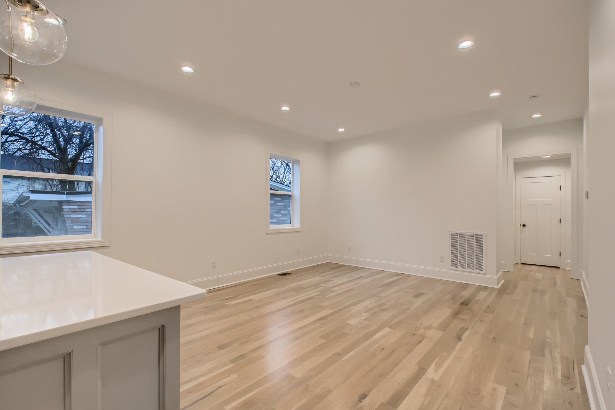 Open living area with wood floors, recessed ceiling lights, glass pendant fixtures and windows along the wall