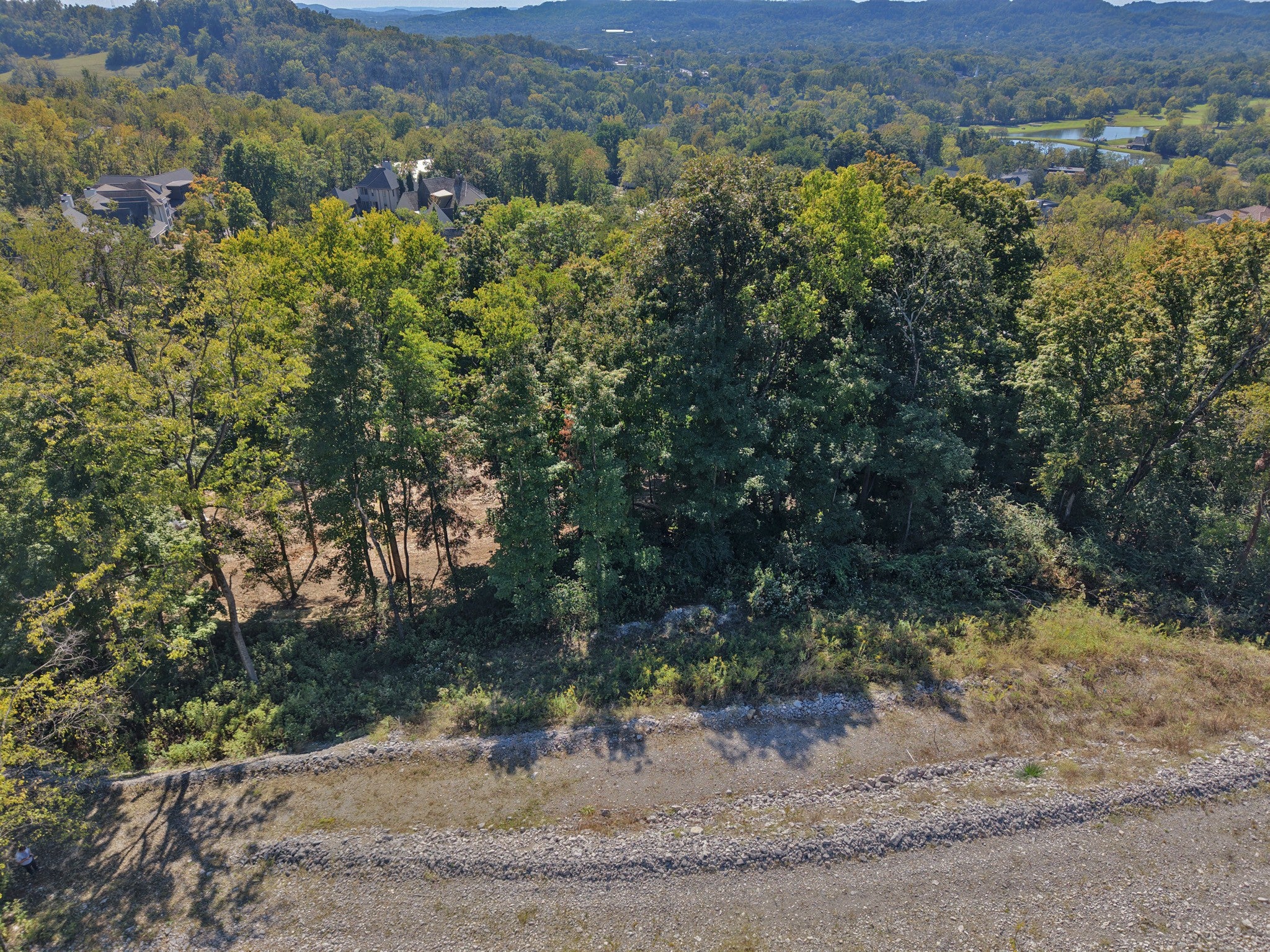 lot for sale in Brentwood, TN 37027, 1765 Woodsong Dr Aerial view of a wooded hillside with a cleared strip along a gravel edge, with rooftops and a valley in the distance
