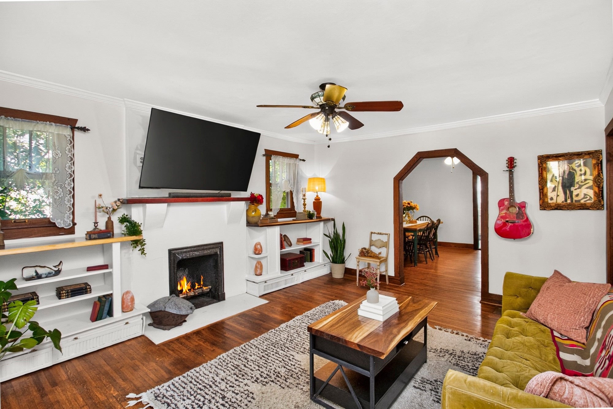 Living room with a white fireplace and built-in shelves, wood floors, a ceiling fan and an arched opening to the dining room