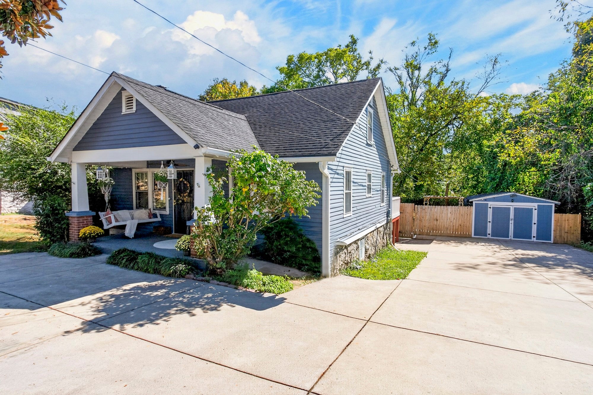 Exterior side view of a gray-blue house with a gabled porch, paved driveway and a blue storage shed behind a wood fence