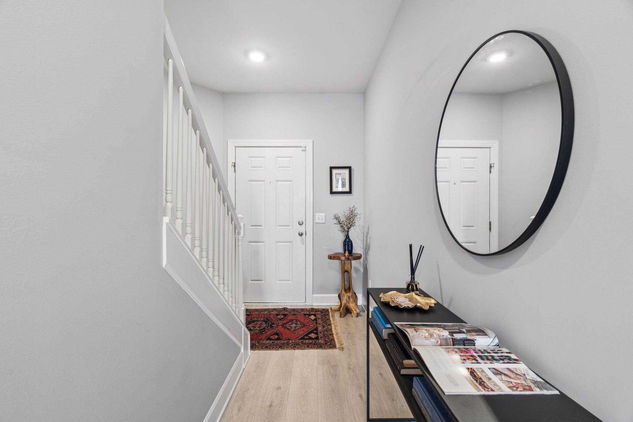 Hallway with wood-look flooring, white six-panel door, a round mirror over a black console table, and a red rug