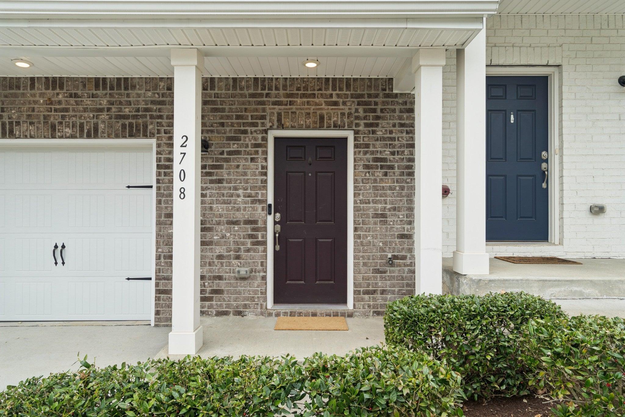 Covered front entry with brick columns, a dark wood front door and white garage door beside it