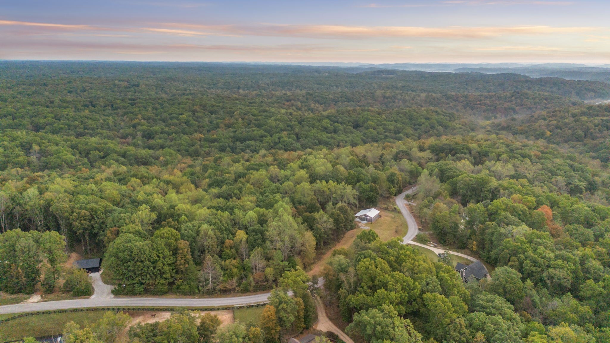 Aerial view over rolling wooded hills with a curving road leading to a barn on a clearing