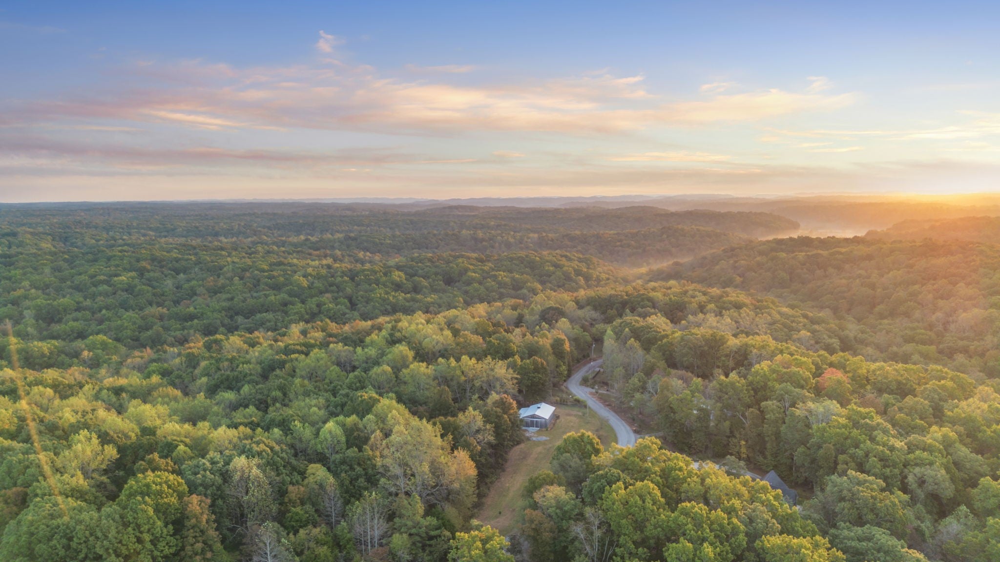 Aerial view at sunset of a barn on a cleared lot within a wide forest of rolling hills