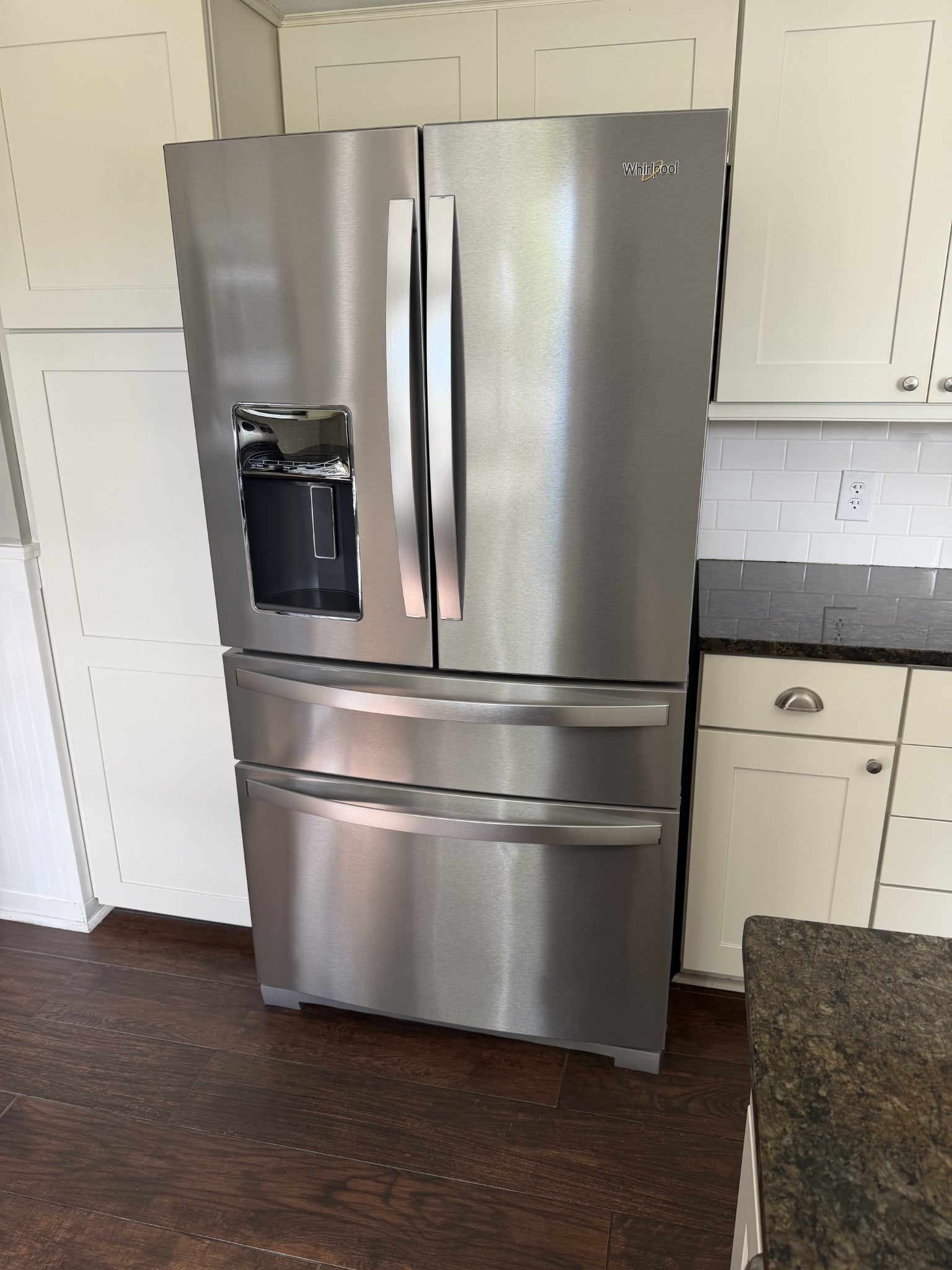 Stainless steel refrigerator with a water dispenser beside white cabinets and dark countertops