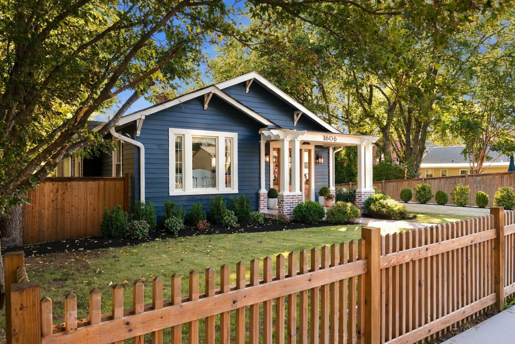 Side view of a blue craftsman home with a window grouping, a covered porch and a wood picket fence in the foreground