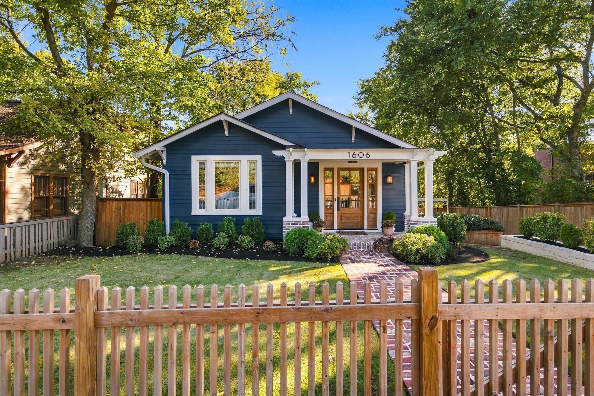 Blue house with a brick walkway leading to a wood front door flanked by sidelights, with boxwood shrubs and a picket fence