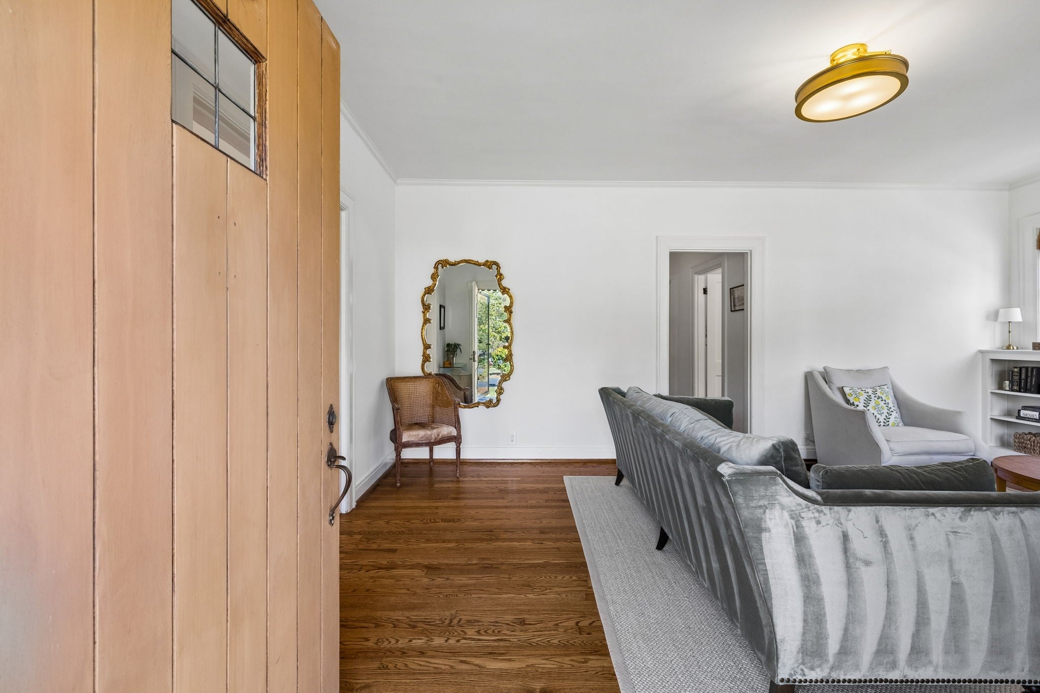 Living room with wood floors, a gold flush-mount light, a gilt mirror and a gray velvet sofa near the front door