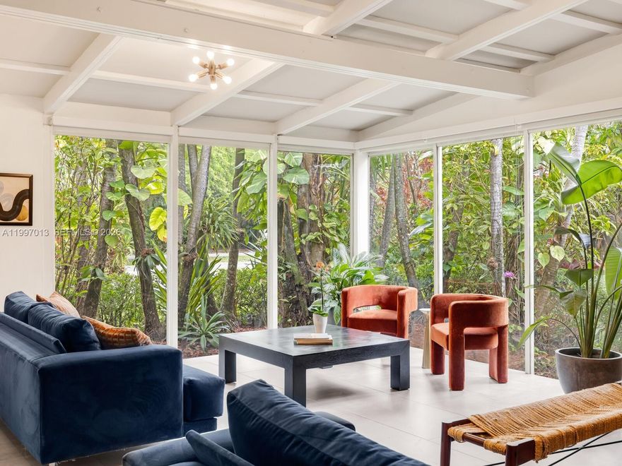 Welcome to Casa Selva in Biscayne Park— a tucked-away tropical mid-century retreat where architecture and nature exist in perfect harmony. Set on an expansive 11,475 SF lot, this 3-bedroom, 2-bath residence offers 2,242 SF of living space enveloped in lush, private gardens. Enter into a dramatic open living, dining, and media space defined by soaring vaulted wood beam ceilings and walls of glass that frame the tropical landscape—creating a serene, gallery-like atmosphere that feels torn from the pages of Architectural Digest. The updated chef’s kitchen features a gas range, eat-in island, and abundant storage. The split floor plan places the primary suite with double-vanity renovated bath on the north side, while two guest bedrooms share a stylish Jack & Jill bath to the south. A spacious Florida room with brick accents opens to a backyard oasis complete with entertaining patio and mango trees. Additional features include new A/C, one-car garage, utility storage, and curated landscape lighting. Hidden, soulful, and architecturally rich—this is Biscayne Park living at its most distinctive.