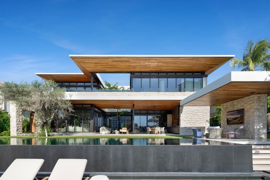 Glamour in Golden Beach! Masterfully designed, this 2025 residence sets a new benchmark in waterfront modernism. Hand-placed stone, iroko wood, and clear glass interlace a 100-year olive tree.  The grand foyer achieves 24' heights highlighting Italian oak-paneled walls that lead to a sophisticated salon. Curated by Fanny Haim, the worldly interiors include a travertine bar in the bespoke media lounge, and stylized fireplaces & drapery throughout. The exceptional MiaCucina kitchen revolves around a bright breakfast area. An architectural staircase arrives to the primary suite’s dual dressing rooms and spacious marble bath enhanced by an upstairs library. A theater, office, gym, elevator & staff room complete the home. The resort pool shines with a cabana, Summer Kitchen, new dock + seawall.