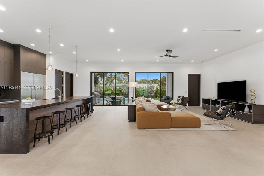 A newly completed 2025 residence by Eitan Construction, this 4-bedroom, 3.5-bath home brings a modern, loft-like sensibility to Wilton Manors. Set on a corner lot, the home offers a sense of openness, privacy, and strong architectural presence. The interiors are defined by scale and natural light, with 11-foot ceilings and oversized ES impact windows throughout. A warm, modern palette of soft grays, deep tones, wood, and bronze accents creates a calm, cohesive living environment. The open living and dining areas center around a thoughtfully designed kitchen, featuring a 13-foot Caesarstone island, custom European cabinetry, a full Thermador appliance suite, and a concealed walk-in pantry. Clean lines and integrated finishes give the space a seamless, uncluttered feel.
Large-format 48x48 porcelain tile floors run throughout, reinforcing the home’s open, continuous layout. Sliding glass doors connect the interior to a covered lanai with a motorized SPF screen, extending the living space outward.

Additional details include Restoration Hardware fixtures, custom closets, and motorized blinds by The Shade Company. Outside, a saltwater pool and spa sit within fully fenced, landscaped, and irrigated grounds, offering a private and easy outdoor setting. Built with long-term durability in mind, the home includes cement slab walls, industrial-grade roof construction, dual-zone Nest climate control, Cooper & Hunter HVAC, Rheem water heating, and a 22x20 two-car garage.