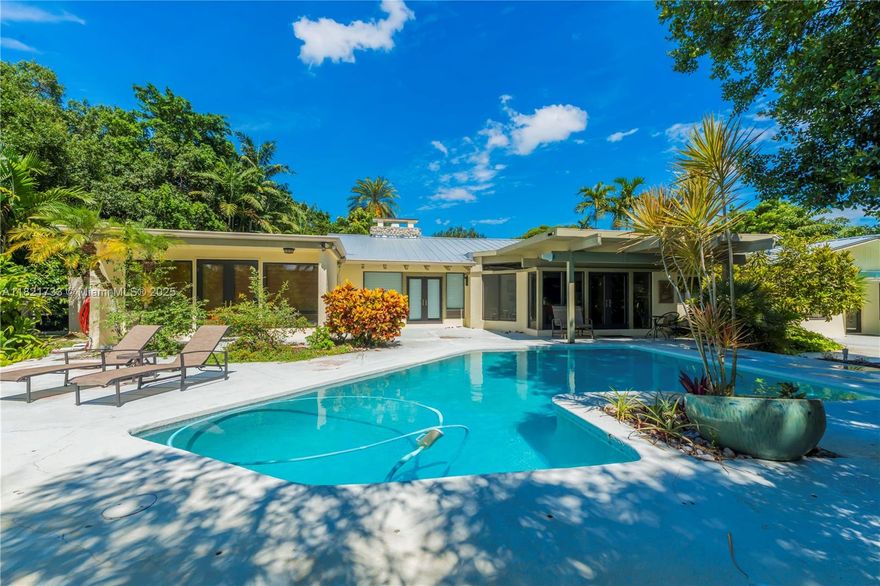 Experience serenity at the edge of the Everglades in this mid-century modern masterpiece. Designed by renowned architect Dan Duckham, a student of Frank Lloyd Wright, this organic home blends wood, coral rock, and expansive glass to bring the outdoors inside. The house features floor-to-ceiling windows and doors along the lakefront, offering stunning views of a fishpond, lush foliage, and tropical fruit trees like key limes and Meyer lemons. Inside, a coral rock fireplace connects the living and family rooms. This 5-bed, 3-bath home boasts recent updates including a new roof, pool resurfacing, impact windows, and modern comforts, making it a unique tropical sanctuary. Some kitchen walls were recolored using AI technology.