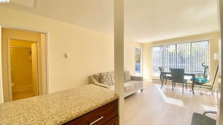Open-concept living area featuring light wood-finish flooring, a large window with vertical blinds, and access to an outdoor balcony