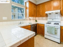 Kitchen with white gas stove, brown cabinetry, tile countertops, and under cabinet range hood