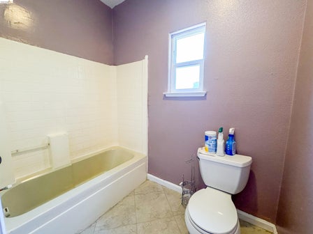 Bathroom featuring a white bathtub with subway tile surround, neutral-toned floor tiling, a white toilet, and a window