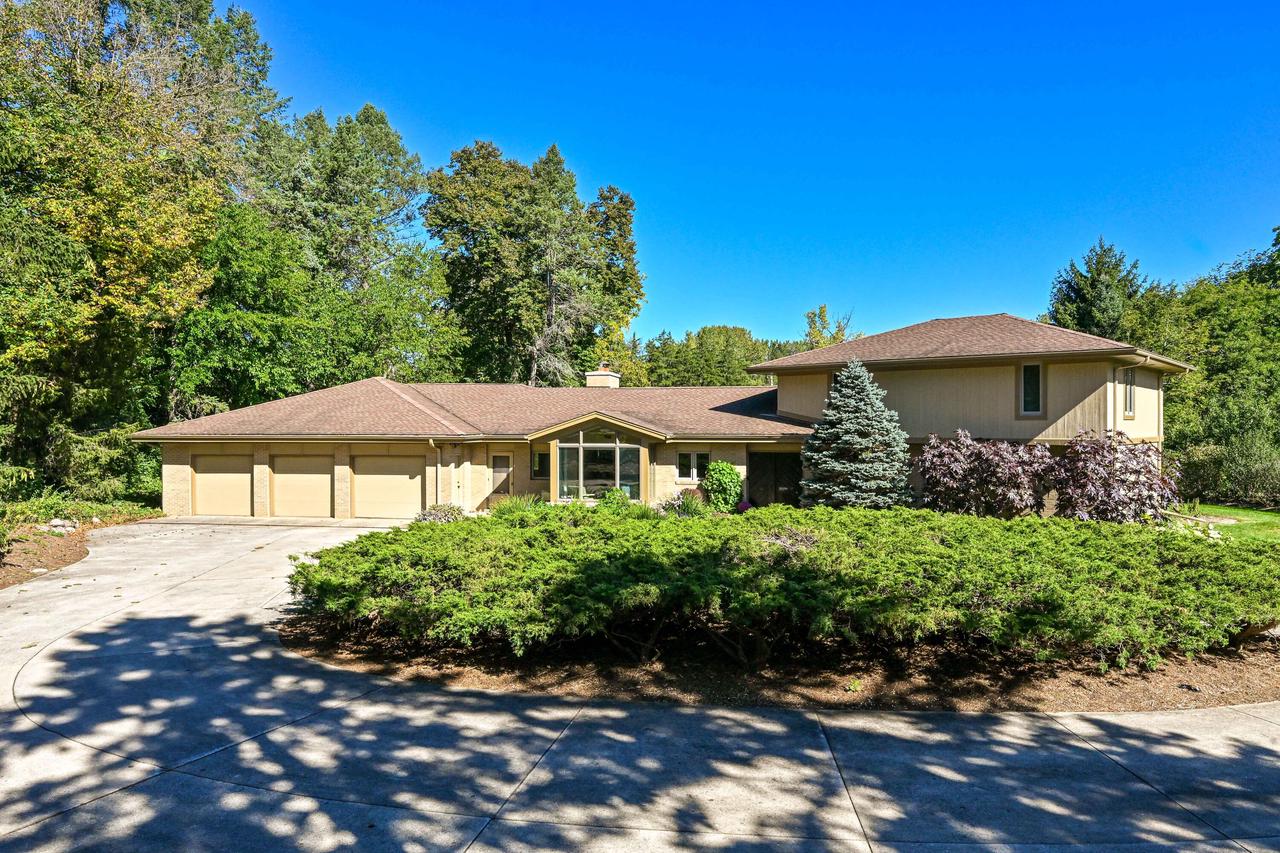 Tan home with a three-car garage, landscaped shrubs, and mature trees.
