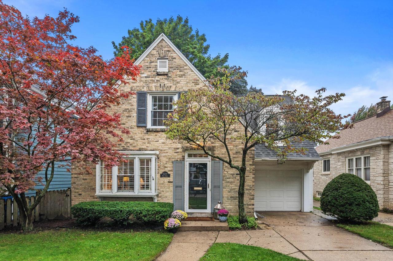 Two-story stone house with a steep gable, attached garage, and red autumn trees.