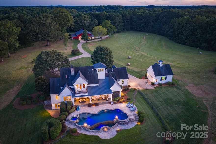 Set across 160 acres in Statesville, North Carolina, this private legacy estate offers a rare opportunity to steward a sanctuary where architecture, agriculture and nature exist in balance. Enter through a winding concrete drive framed by mature hardwoods, a producing orchard and vineyard, and arrive at a 6,617-square-foot residence custom designed to embrace the land’s grandeur while offering every modern comfort.

Built in collaboration with a respected architect, builder and interior designer, the residence spans three finished levels plus a tower room providing panoramic sunset views over the Blue Ridge foothills. An expansive covered porch welcomes guests into a gracious main level anchored by a great room with fireplace, an epicurean kitchen, breakfast room and keeping room — each with access to a multi-level veranda and pool terrace. Custom cabinetry, dual islands, top-tier appliances and a scullery define the kitchen’s function and elegance. A screened porch with a two-sided fireplace encourages seamless indoor-outdoor living.

The primary suite offers long views of open fields and forest, a spa-caliber bath and custom built-in cabinetry in large walk-in closet. A secondary guest suite is positioned for privacy on the main floor. The west wing includes a mudroom, laundry, powder room, office and access to an oversized side-entry two-car garage.

Upstairs, two en-suite guest bedrooms with window seats, a loft library with built-ins, two full baths and an office or the 5th bedroom.  A flex room/music room provide additional living and working space. The tower room, accessed from the second level, presents unmatched views across the estate’s wooded canopy and surrounding mountain horizon.

The lower level includes a lounge/sitting and game room with wet bar and wine cellar, a fitness studio, full bath, flex room, cedar-lined storage and unfinished mechanical space.

Beyond the residence, a motor court links to a detached two-car carriage house garage. Nearby, a 3,000-square-foot workshop includes three 12-foot motorized doors, a concrete floor, kitchenette, full office suite with HVAC and bath, a loft and a full-length lean-to for storage or agricultural use. Designed for versatility, this structure accommodates luxury vehicle storage, equipment, creative studio or event conversion.

Zoned for agriculture and recreation, the property offers long-term land use flexibility and is ideally suited for multigenerational living, equestrian development or conservation easement potential.

Hay fields, woods, creek and privacy all located less than 10 minute to I-77 and I-40 Junction and Statesville.  Easy access to Lake Norman, Winston Salem, Hickory and Charlotte.