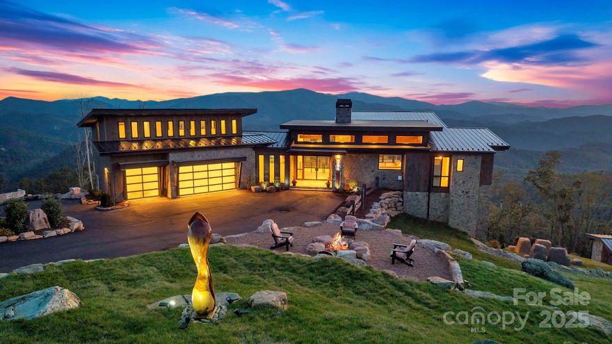 Located in the coveted Sunset Section of The Lodges at Eagles Nest, the Eagles Nest Glass House is an exceptional custom estate redefining mountain living through masterful design and craftsmanship. Developed over three years, this architectural showcase occupies 1.033 acres, where rugged natural beauty blends seamlessly with refined elegance. The residence offers 7,055 sq ft of heated living space, excluding the three-car garage. Perched at 4,400 feet elevation, it commands panoramic views spanning North Carolina to Tennessee mountains, framed by awe-inspiring sunsets. The exterior presents a stylish mountain retreat with white pine-stained wood siding, expansive windows, and rustic metal beams. Over $250,000 was invested in professional landscaping, featuring natural boulders, enhanced lighting, and intricate hardscaping that create an impressive arrival experience. Outdoor living shines with 860+ sq ft of decking, a large hot tub, hand-laid stone pillars, and custom steel beams designed to highlight the sweeping vistas. An inviting fire pit promises memorable evenings beneath the stars. Entry by a grand custom glass door reveals a great room with soaring two-story glass walls that flood the interior with natural light while framing the stunning surroundings. Nickel gap white pine-stained ceilings with rustic beams combine rustic warmth and modern elegance, accented by hand-laid rock, light-toned floors, and a custom rusted gas fireplace. The gourmet kitchen impresses with Onyx Waterfall Island countertops, stainless steel appliances, and a wine cooler. The lower level houses a large custom bar, wine cellar, and a flexible bonus room suited for theater, family gatherings, or games. Throughout are handmade floating handmade stairs, custom wood ceilings, stone accents, custom glass windows, and exterior stonework. There are five spacious bedrooms, each with meticulously finished bathrooms featuring hand-laid stone, marble, various tiles, and teakwood benches. Two additional half-baths are located on each floor. Two generous primary suites, one on the main level and one on the lower level, feature spa-inspired bathrooms and conveniently located adjacent utility rooms. Above the garage, featuring hand painted bronze epoxy floors, lies a distinctive bunk room with a full bath and an expansive rec room. Roughly half of this 275 sq ft area features ceiling heights between 5 and 7 feet. The home is enhanced by commissioned artwork and custom furnishings throughout, reinforcing its one-of-a-kind character. Residents enjoy an extensive amenity offering including ATV and hiking trails, ropes course, playground, dog park, mini-golf, disc golf, archery, versatile sports fields, and courts for volleyball, basketball, tennis, and pickleball. Enjoy world class trout fishing along the Elk River with over 2000 ft of river frontage. Dining options include Eagles Nest Winery, Epic Chophouse and The Sportsman’s Grille. Additional amenities include a 24-hour Dollar General, an outdoor amphitheater offering live entertainment on Saturday evenings from May through October, fitness center, and two helicopter pads. This estate transcends standard mountain homes by flawlessly combining luxury, nature, and architectural brilliance, inviting an elevated mountain lifestyle. 4 year home warranty provided