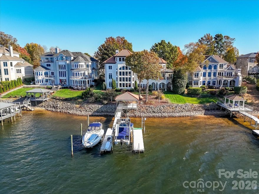 Architecturally distinctive Mediterranean-inspired waterfront estate by renowned builder G.M. Helms, thoughtfully designed in collaboration with architect Christopher Phelps and celebrated interior designer Dina Lowery. Featured on the cover of Charlotte Magazine as an architectural wonder, this four-story residence is ideally positioned on Lake Norman’s main channel with west-facing exposure and captivating sunset views.
Designed to be both grand and inviting, the home unfolds through a series of refined yet comfortable spaces. A dramatic three-story great room anchors the residence, showcasing soaring ceilings, a monumental limestone fireplace, and an expansive wall of lake-facing windows that flood the home with natural light. A striking four-story, self-supported circular staircase winds gracefully through all levels, serving as the home’s defining architectural focal point.
The residence offers approximately 7,400 square feet of living space, including five bedrooms, eight baths, six fireplaces, a Brazilian cherry wood library, media room, billiards room, and an elevator serving the first three levels. Designed with entertaining in mind, the home features a chef’s kitchen on the main level and a full secondary kitchen on the lake level, along with multiple gathering and leisure spaces throughout.
The fourth-floor sky lounge with bar provides elevated views of the lake and surrounding islands, creating a unique retreat for entertaining or quiet relaxation. Outdoor living is equally impressive, with three balconies, a Spanish-tiled lakeside gazebo, and a deep-water dock equipped with lifts for two jet skis, a double-decker pontoon, and pilings suitable for a 45-foot yacht.
Lovingly maintained by only two owners since its 1997 construction, the home has been extensively updated, including renovated kitchens and baths, four HVAC systems, two tankless water heaters, enhanced audiovisual systems, exterior and interior repainting, and refreshed landscaping. A rare offering that seamlessly blends timeless Mediterranean design, thoughtful craftsmanship, and premier Lake Norman waterfront living.