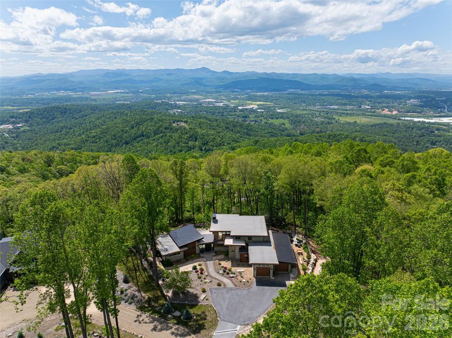 Embrace refined mountain living in this striking contemporary estate, where serenity and sophistication converge atop a rare flat mountaintop setting in the gated conservation community of Couch Mountain. Sweeping 180-degree Blue Ridge Mountain views unfold through expansive walls of glass, creating a breathtaking backdrop from nearly every room while timeless craftsmanship and elevated design define the living experience.

This exceptional property is being offered three separate ways to suit a variety of lifestyles and investment opportunities: this listing includes the residence and the individual lot it sits on; the home is also available with both the individual lot and the adjoining lot; and the additional homesite may be purchased separately. Together, the offerings present a rare opportunity to secure privacy, flexibility, and long-term value in one of Western North Carolina’s most distinctive mountain communities.

Inside, the dramatic great room showcases soaring 20-foot ceilings, hand-hewn 125-year-old beams reclaimed from a Canadian barn, exposed custom steel supports, and a stunning stacked-stone fireplace anchored by a concrete hearth. Designed for both grand entertaining and comfortable everyday living, the open-concept floor plan flows effortlessly into the gourmet kitchen, where Thermador stainless steel appliances, Italian quartzite countertops, and bespoke rustic alder cabinetry create a space that is as functional as it is beautiful. A walk-in pantry and coffee bar, concealed behind a sliding knotty alder barn door, add warmth and character.

The adjoining dining area and sunroom—with its inviting stone fireplace and panoramic mountain vistas—offer seamless indoor-outdoor living and memorable gathering spaces year-round. The main-level primary suite is a luxurious retreat featuring breathtaking views, a spa-inspired oversized shower, and a custom walk-in closet thoughtfully designed for comfort and organization. Upstairs, two private ensuite guest bedrooms showcase artisanal tilework and refined finishes, providing elevated accommodations for family and visitors alike.

Outdoors, the curated mountain landscape invite exploration and relaxation with established gardens, walking trails, natural stone features, and a fenced vegetable garden. Whether enjoying sunrise coffee on the terrace, entertaining beneath the stars, or simply taking in the stillness of the mountains, the setting is both peaceful and inspiring.

Ideally located just 15 minutes from Asheville Regional Airport and conveniently positioned between Asheville and Hendersonville, the property offers easy access to acclaimed restaurants, shopping, arts, and outdoor recreation. Community-approved seven-day rentals also create the opportunity for a lock-and-leave lifestyle with potential income-producing flexibility when not in residence. Rarely does a property combine architectural distinction, panoramic views, privacy, and versatility so seamlessly in one extraordinary mountain offering.
