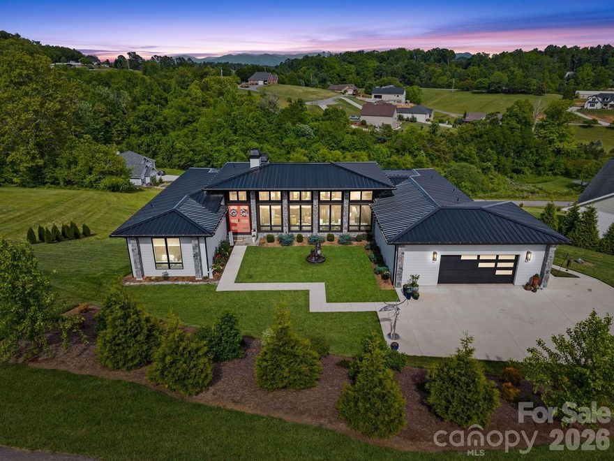 A rare offering in the heart of Western North Carolina, this distinguished custom residence was featured in the 2023 WNC Parade of Homes and represents the pinnacle of modern luxury living. Crafted by Dwelling Place Homes and aptly named "The Ridge," the home commands its setting with effortless authority — where masterful architecture, impeccable craftsmanship, and an idyllic natural backdrop converge in perfect harmony.
Situated on a coveted double lot spanning 1.51 acres within an exclusive gated community, this property offers a level of privacy and prestige that is truly unmatched. Residents enjoy access to resort-style amenities including a community pool, pavilion, and basketball court — an exceptional lifestyle offering rarely found at this caliber.
The approach alone is unforgettable. Natural stone columns frame walls of soaring black-framed glass, while a bold coral entry door and sculptural courtyard fountain signal the artistry within. Once inside, 13-foot ceilings and an abundance of natural light create an atmosphere of serene openness, anchored by a dramatic floor-to-ceiling polished stone-look porcelain fireplace that serves as the room's undeniable focal point.
The culinary suite is a study in refinement. Conceived by the celebrated Expressions Cabinetry, it showcases custom cabinetry, breathtaking book-matched marble countertops, a professional Z-Line gas range, built-in Miele coffee system, beverage cooler, and a luminous marble tile backsplash — a space as beautiful to behold as it is to cook in.
The outdoor living experience is equally impressive. A generous composite deck with outdoor fireplace and cable railing offers an unparalleled setting for both intimate evenings and grand entertaining under the Carolina sky.
Finished with a standing seam metal roof, smart siding, natural stone exterior accents, and lush professional landscaping, every element of this home has been considered with intention. Opportunities of this distinction are exceedingly rare — this is Western North Carolina living at its most extraordinary.