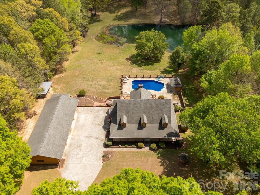 Welcome to a truly rare luxury estate set on over 3 acres, offering more than 5,600 heated square feet designed for those who crave space, privacy, and a resort-style lifestyle. Ideally located just minutes from Charlotte, this exceptional property offers the perfect balance of convenience and seclusion, creating a true private retreat.

Tucked into a peaceful setting, this custom wood-sided home makes an unforgettable first impression with its long driveway, manicured grounds, and inviting wraparound front porch—the perfect place to slow down and take in the surroundings, where it’s not uncommon to spot wild turkeys wandering through the backyard.

Inside, the home opens to a stunning two-story great room anchored by a statement fireplace and framed by double French doors that lead to an expansive back deck. From here, the view unfolds into a private outdoor oasis featuring a sparkling in-ground pool with a diving board and slide, along with a stocked pond that enhances the serene, getaway-like atmosphere.

The main level is thoughtfully designed for both comfort and flexibility, featuring a spacious primary suite that sets a new standard for relaxation with its spa-like bath, complete with a soaking tub, dual granite vanities, built-ins, a large walk-in ceramic shower, and the added luxury of two walk-in closets. The main floor also includes a well-appointed kitchen and a flexible bedroom or office space, making it ideal for remote work or guests.

Upstairs, two additional bedrooms, a full bath, and a loft overlook the pool and pond, creating a picturesque setting for a second living area, reading nook, or media space.

The walk-out basement is perfectly suited for extended living or entertaining, offering an expansive recreation room with built-in trophy and display cases, a wet bar, billiards area, fireplace, half bath, and a dedicated workout room. With direct access to the pool deck, this level provides excellent potential for multi-generational living, guest quarters, or a private retreat within the home.

Outdoors, the property continues to impress with a large back deck, a beautifully maintained fenced yard, and carefully curated landscaping accented by brick columns, hardscaping, and wrought iron details. Additional structures include a pool house, a four-bay detached garage, and dog kennels, providing both functionality and flexibility for a variety of lifestyles.

With walk-in attics, custom built-ins throughout, and every detail designed with intention, this home offers the perfect blend of luxury, space, and versatility. For those seeking a private estate with acreage, a pool, a pond, and room to accommodate multi-generational living—all within minutes of Charlotte—this is a rare opportunity to own a property that truly feels like a destination.