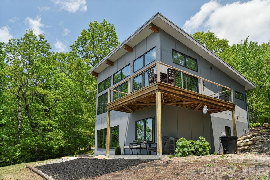 Perched high above the valleys, this striking modern mountain retreat offers a rare blend of sophistication, privacy, and panoramic beauty. Designed to capture some of the region’s most captivating long-range views, the residence frames the landscape through soaring floor-to-ceiling glass, allowing the ever-changing mountain vistas to become a living work of art within your home. The interior unfolds in an open and inviting layout, where contemporary finishes meet timeless comfort. A gas-log fireplace anchors the great room, providing a warm counterpoint to the expansive windows and lofty ceilings. The primary suite, thoughtfully positioned on the main level, affords both convenience and serenity. The lower level reveals a self-contained living space with two additional bedrooms, a full bath, and a secondary gathering room—ideal for extended family, guests, or income-producing rental use. Outdoor living is equally impressive, with a sweeping wraparound deck designed for both quiet reflection and lively gatherings under the stars. Practical features include paved access, an upper circle drive, a lower drive with additional parking and private entry to the lower level, and the option to add a garage should you wish to expand. Nestled within a secure, beautifully maintained gated community, residents enjoy tranquil walking trails and an atmosphere of peace and exclusivity. Currently operated as a highly successful Airbnb, the property presents not only a captivating lifestyle opportunity but also exceptional investment potential. Here, modern comfort and mountain majesty converge—creating a retreat that is as versatile as it is unforgettable.