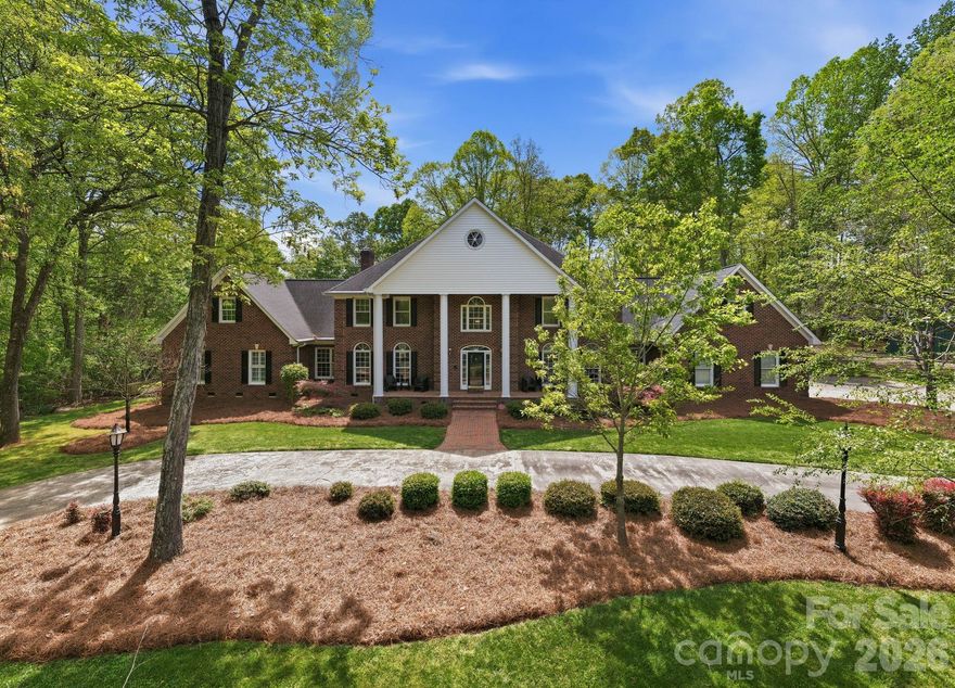 Classic Southern brick estate on 7.11 private wooded acres, just ~35 minutes from Uptown Charlotte and Charlotte Douglas International Airport. A quarter-mile circular driveway winds through mature hardwoods to a stately columned facade. 5 bedrooms, 5.5 baths, and approximately 6,188 sq ft over a thoughtful floor plan with 9-ft ceilings, three staircases, and a two-story foyer. Main-level primary suite with renovated bath (2023). Chef's kitchen renovated 2019 — Wolf cooktop, double ovens, steam oven and microwave drawer, Bosch dishwasher, two 36" refrigeration units, granite, and dual pantries. Upstairs offers four bedrooms, additional finished suite-style space, and a bonus room with new LVP flooring. Outdoors: saltwater pool with full pool house, gas fire pit on a stone terrace, covered grilling porch, and a detached barn studio/gym with mini-split and running water. Three-car attached garage, whole-house natural gas generator (rebuilt 2025), well, septic, and irrigation. Top-rated Clover School District. A rare combination of privacy, scale, and quality — minutes from Lake Wylie's marinas and the conveniences of Charlotte.
